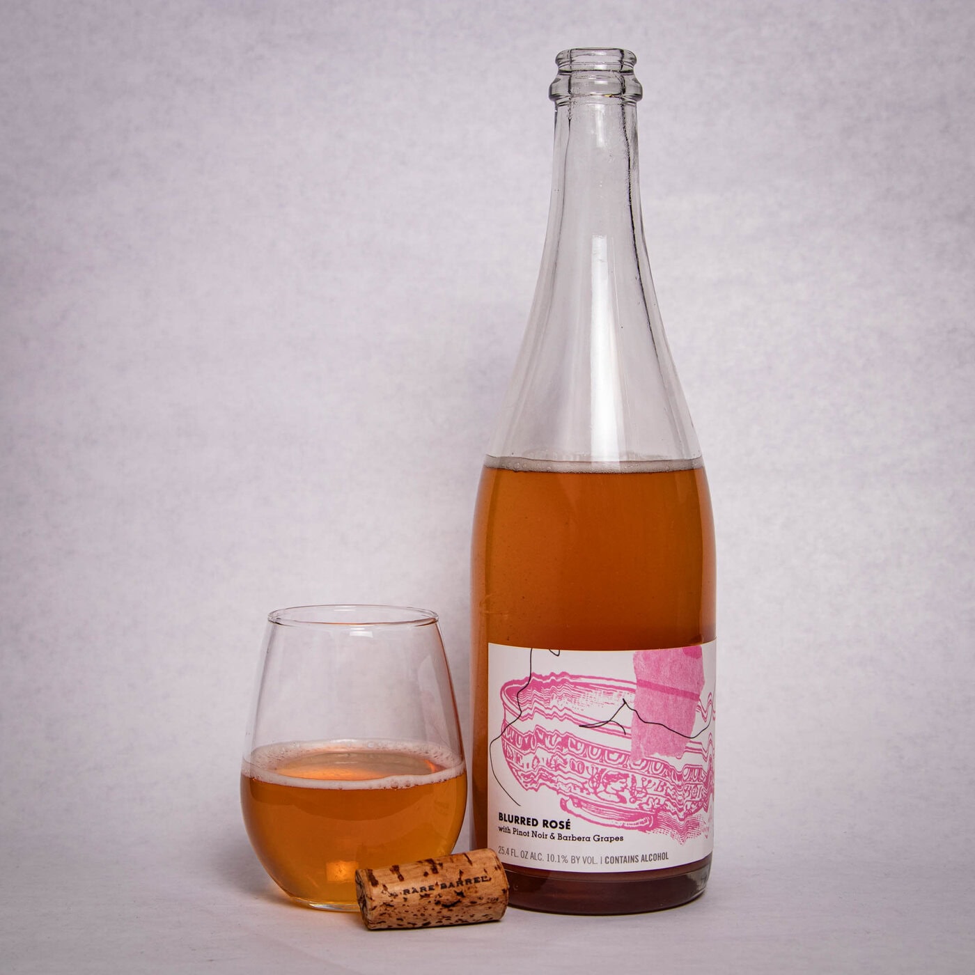 A Clear Wine Bottle With A Pink Illustrated Label Stands Next To A Filled Wine Glass And A Cork On A Light Surface, All Containing A Rose-Gold Colored Liquid.
