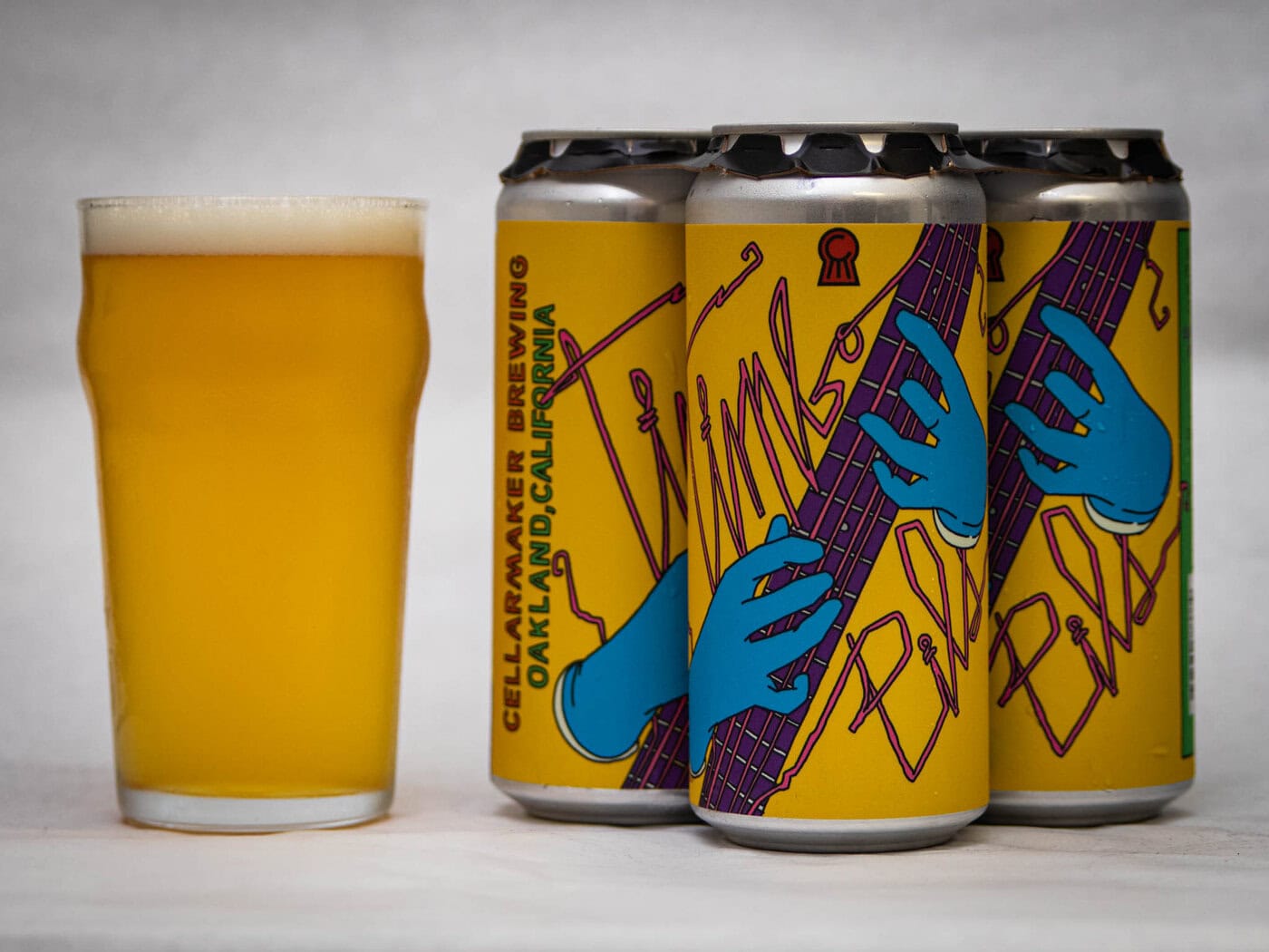 A Pint Glass Of Hazy, Golden Beer Stands Next To Three Cans With Yellow Labels Featuring Blue Hands Playing A Purple Guitar. The Cans Are Labeled &Amp;Quot;Cellarmaker Brewing, Oakland, California.