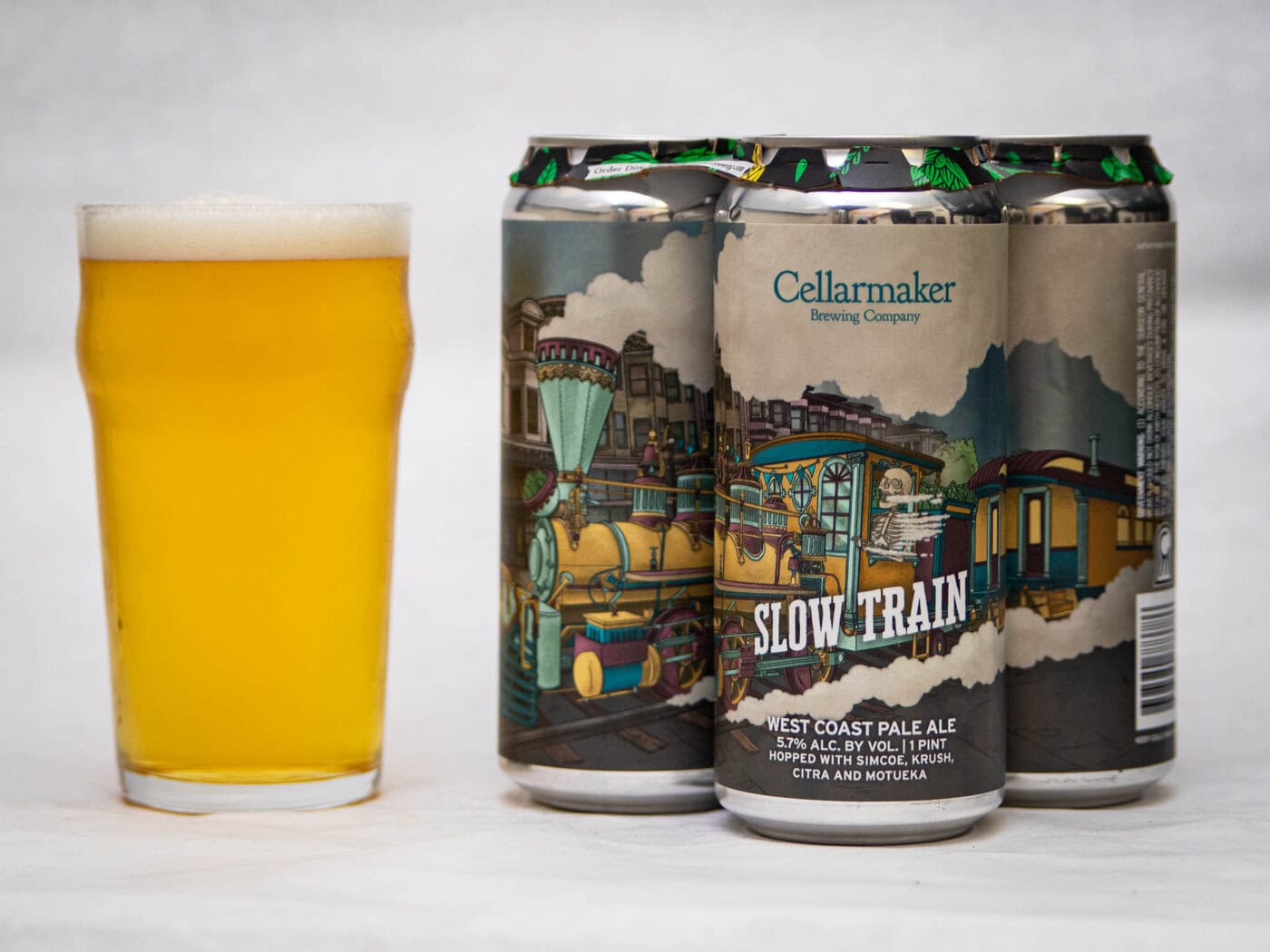 A Hazy Pale Yellow Beer Sits Next To Three Cans Of Slow Train - Wc Pale Ale, Each Featuring Vibrant Train-Themed Artwork On The Label.
