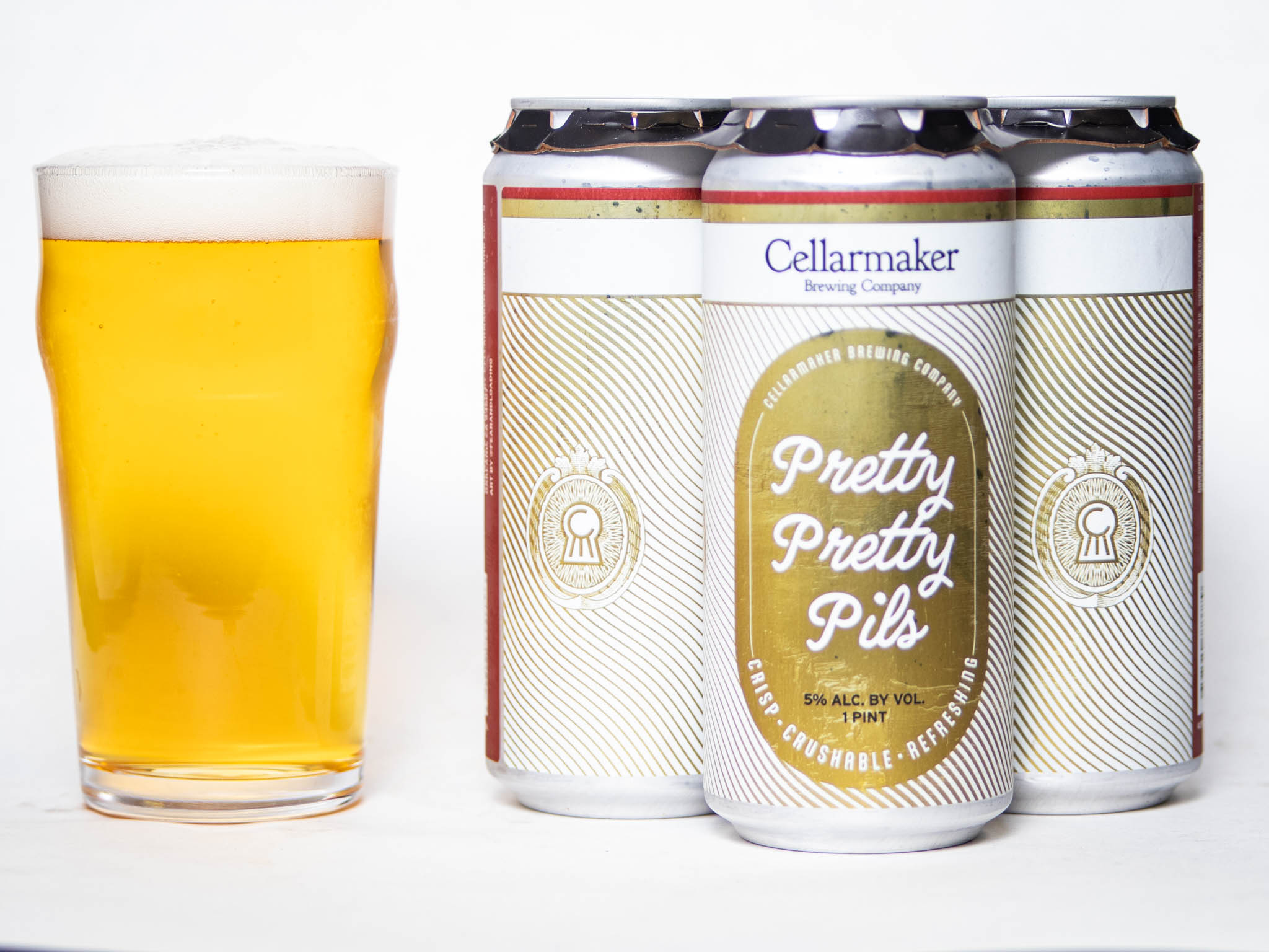 A Pint Glass Of Golden Beer Sits Next To Three Cans Labeled &Amp;Quot;Pretty Pretty Pils&Amp;Quot; From Cellarmaker Brewing Company, With Gold And White Designs On The Cans, All Set Against A White Background.