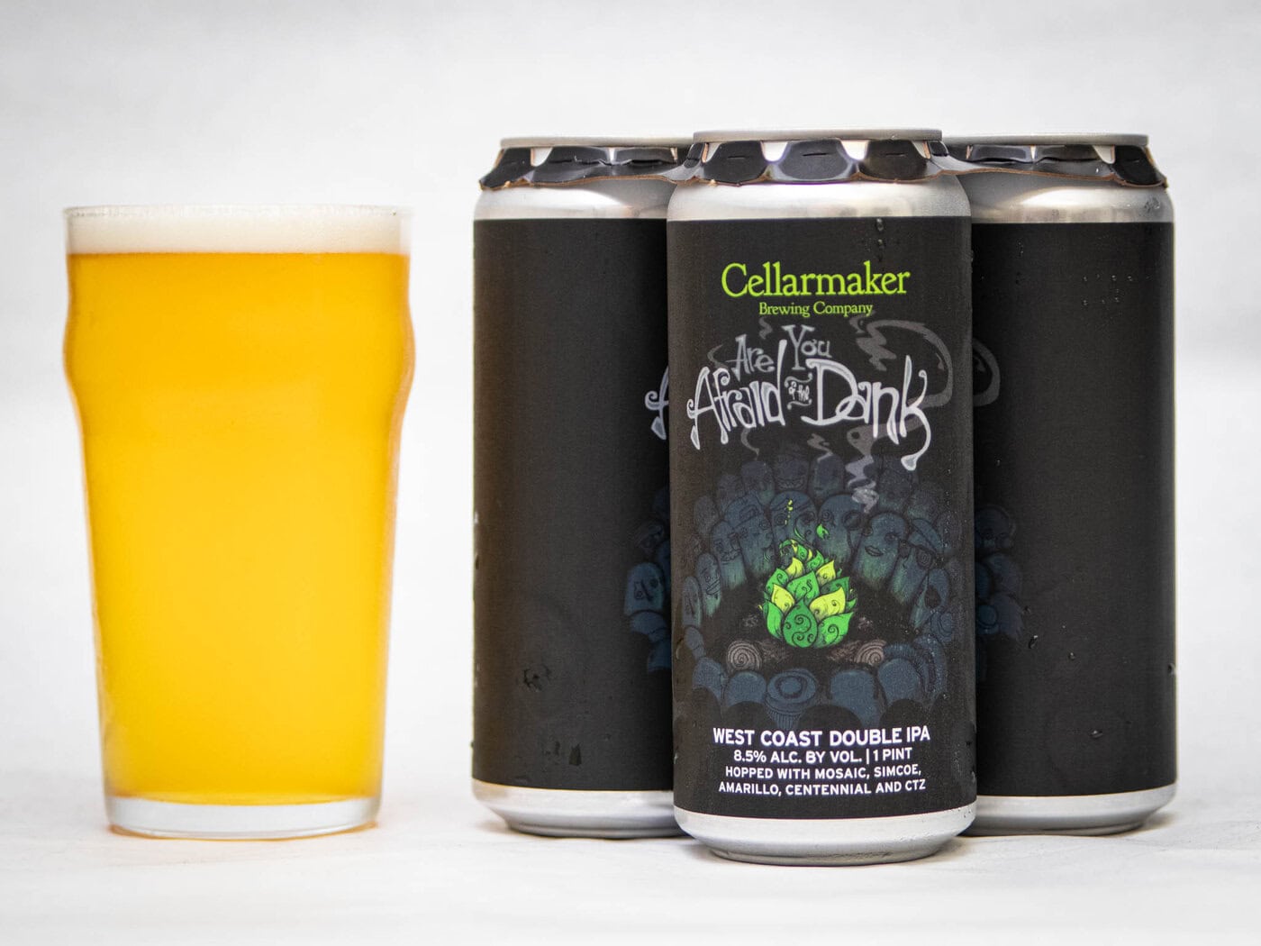 A Four-Pack Of Cellarmaker Brewing Company’s “The Art Of The Dank” West Coast Double Ipa Cans Sits Beside A Full Pint Glass Of Hazy Golden Beer On A White Background.