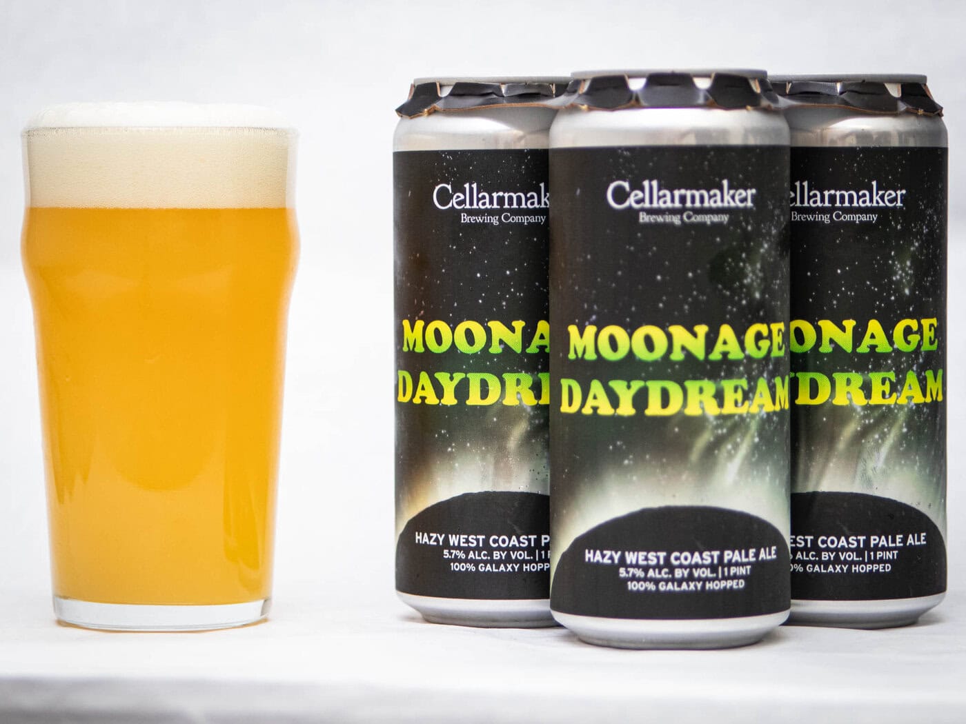 A Pint Glass Of Hazy, Golden Beer Sits Beside Three Cans Of Moonage Daydream - Hazy Pale Ale From Cellarmaker Brewing Company, Each Featuring A Starry Night Design.
