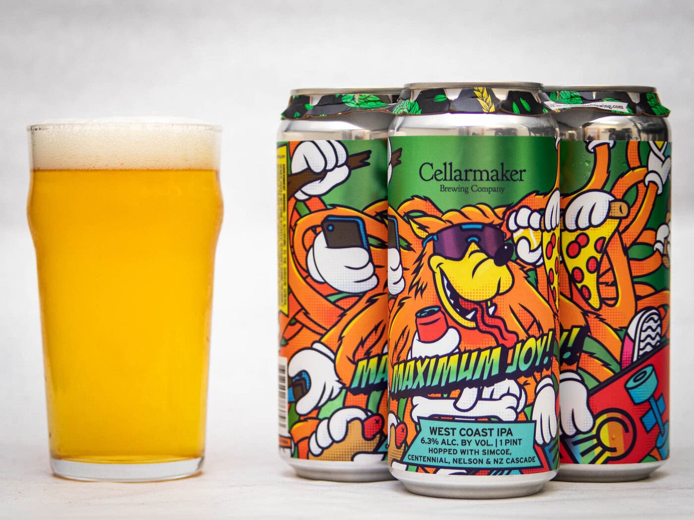 A Pint Glass Of Golden Beer Sits Next To Four Colorful Cans Of Cellarmaker Brewing Company’s Maximum Joy! West Coast Ipa, Featuring Cartoon Eagles And Pizza On The Label With A Vibrant, Playful Design.
