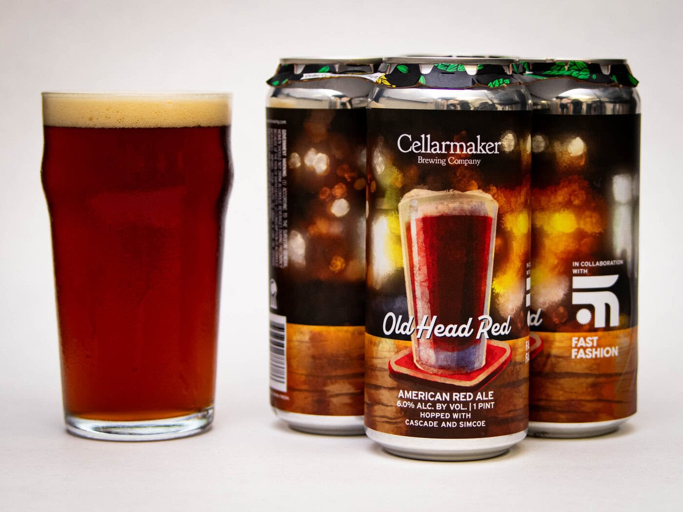 A Pint Glass Filled With Amber-Red Beer Stands Next To Four Cans Labeled “Old Head Red” From Cellarmaker Brewing Company. The Cans Feature A Picture Of The Same Beer And Decorative Bokeh-Style Lights In The Background.
