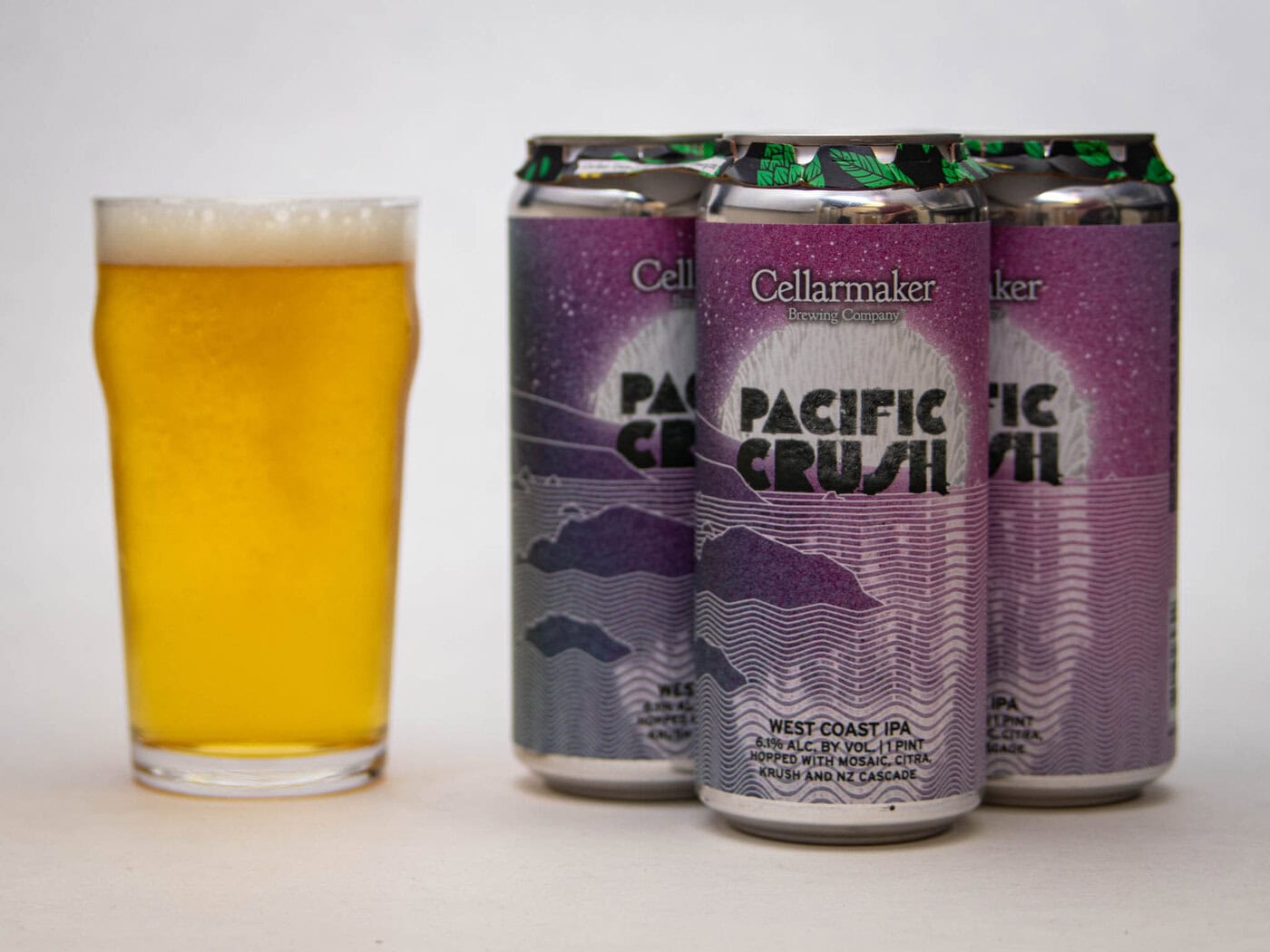 A Pint Glass Filled With A Golden Beer Sits Next To Three Cans Of Cellarmaker Brewing Co.'S Pacific Crush West Coast Ipa, Featuring A Purple And White Design With Waves And Mountains.