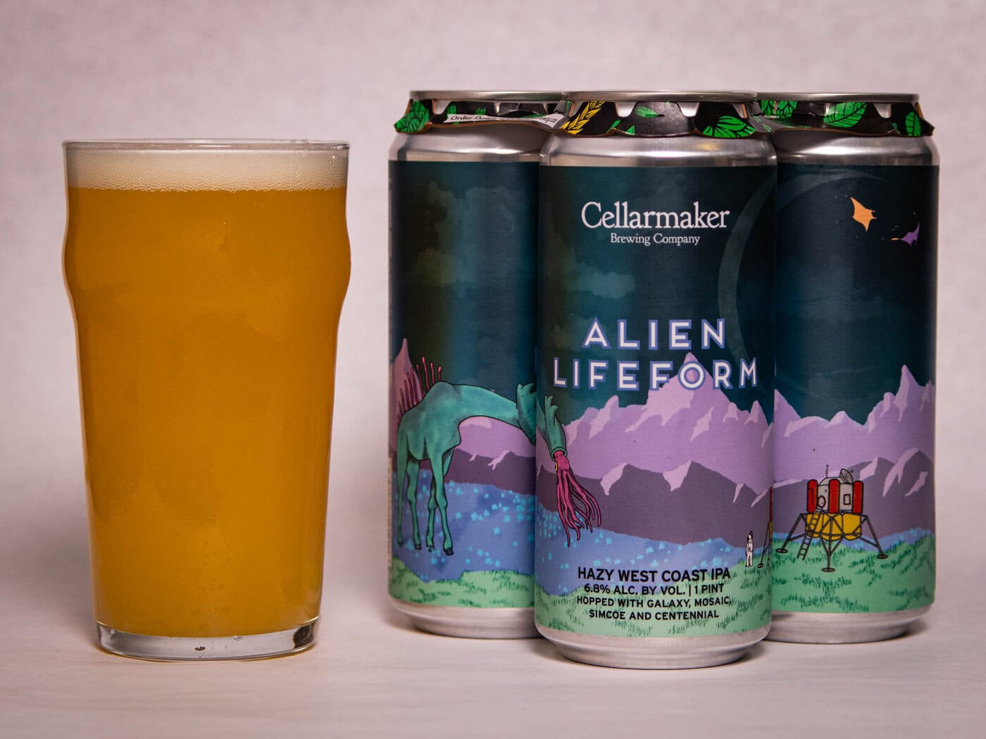 A Pint Glass Filled With Hazy, Golden Beer Sits Next To Three Cans Of Cellarmaker Brewing Company'S &Amp;Quot;Alien Lifeform&Amp;Quot; Ipa, Featuring Colorful Artwork Of An Alien Landscape With A Horse, Mountains, And A Small Rocket.