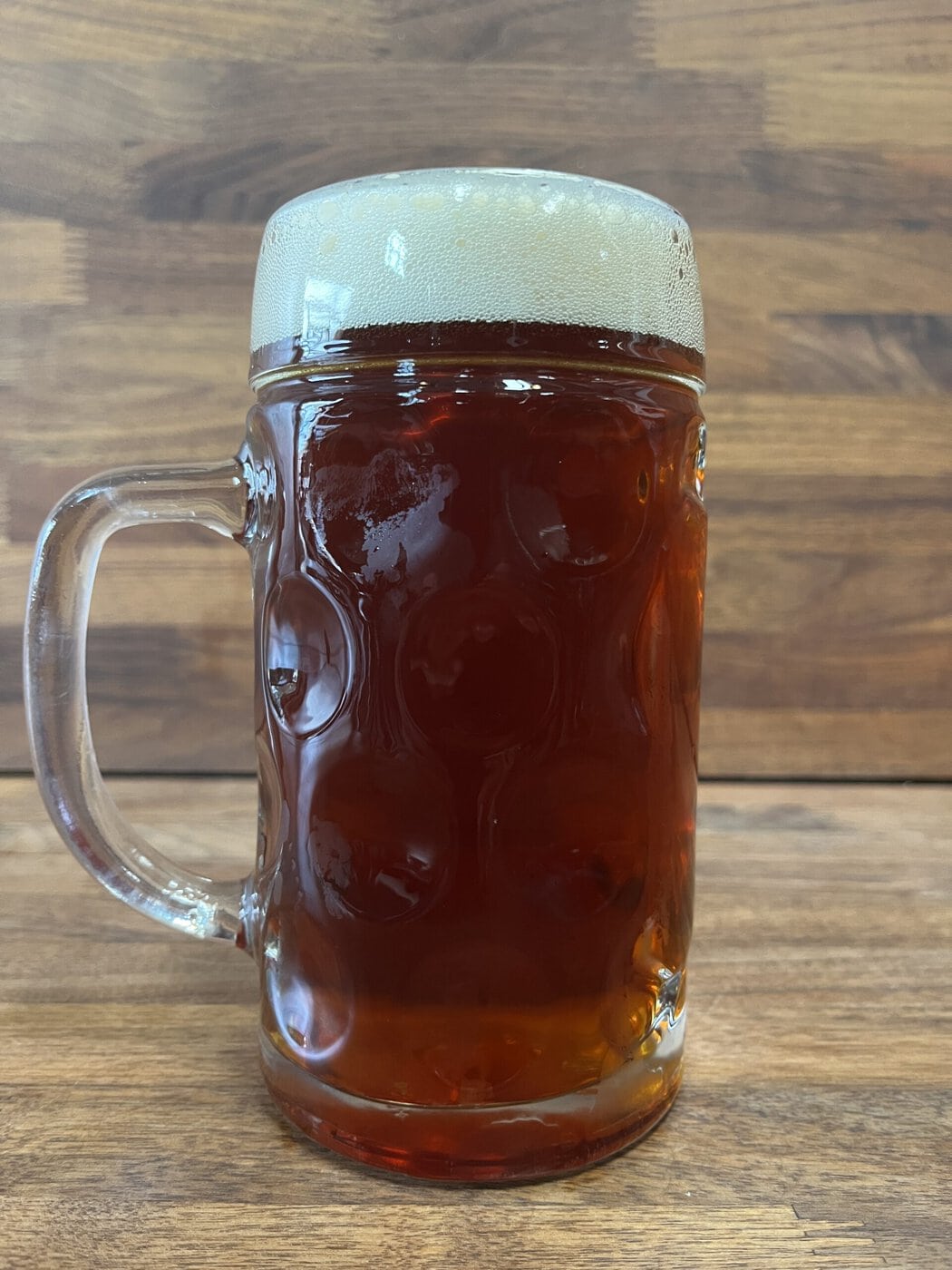 A Large Glass Stein Filled With Dark Amber Beer Topped With A Thick, Frothy Head, Placed On A Wooden Surface With A Wooden Background.