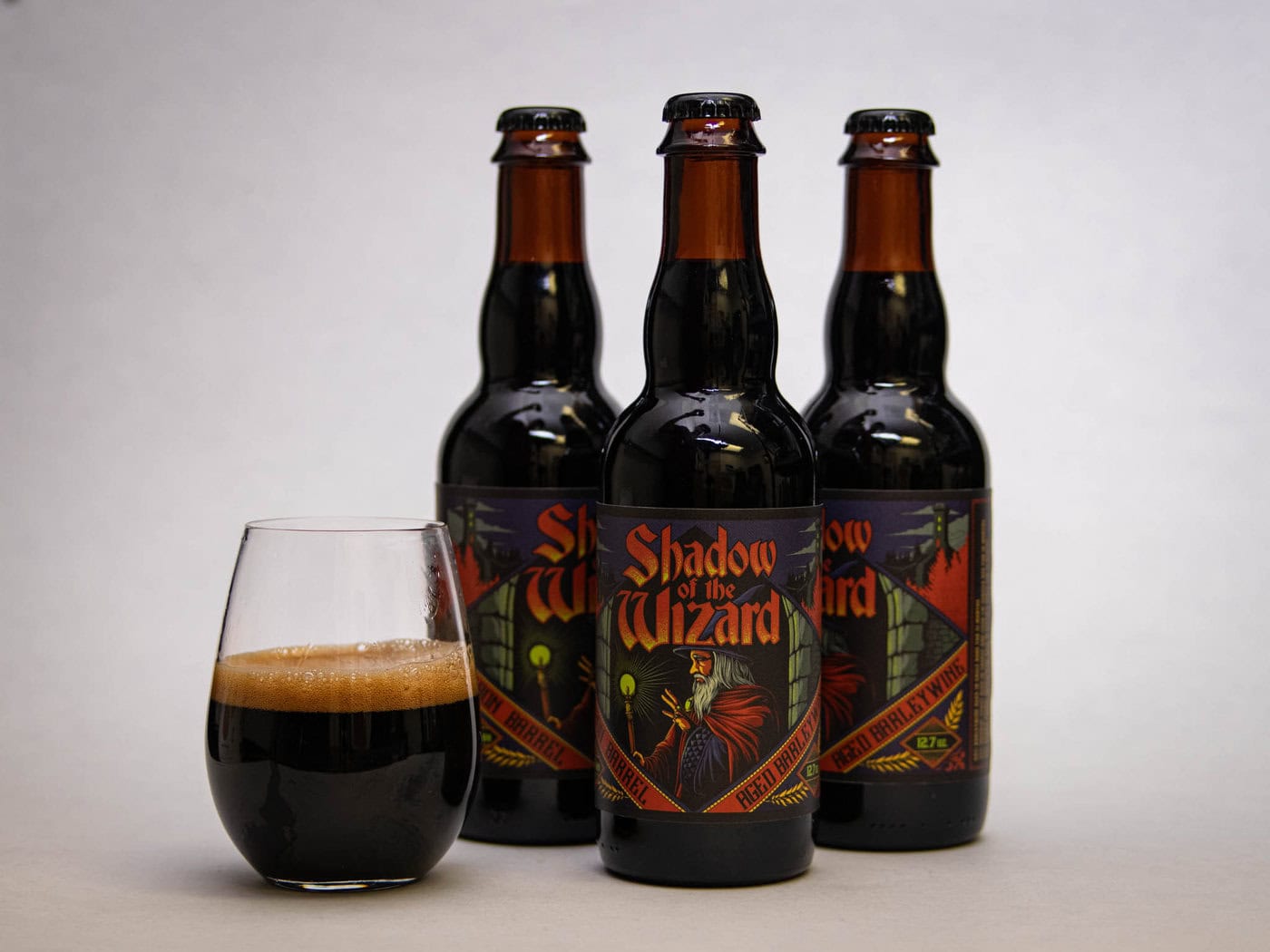 Three Bottles Of &Amp;Quot;Shadow Of The Wizard&Amp;Quot; Beer With Dark Fantasy-Themed Labels Stand Behind A Glass Filled With Dark Beer And A Foamy Head, All Set Against A Plain Light Background.