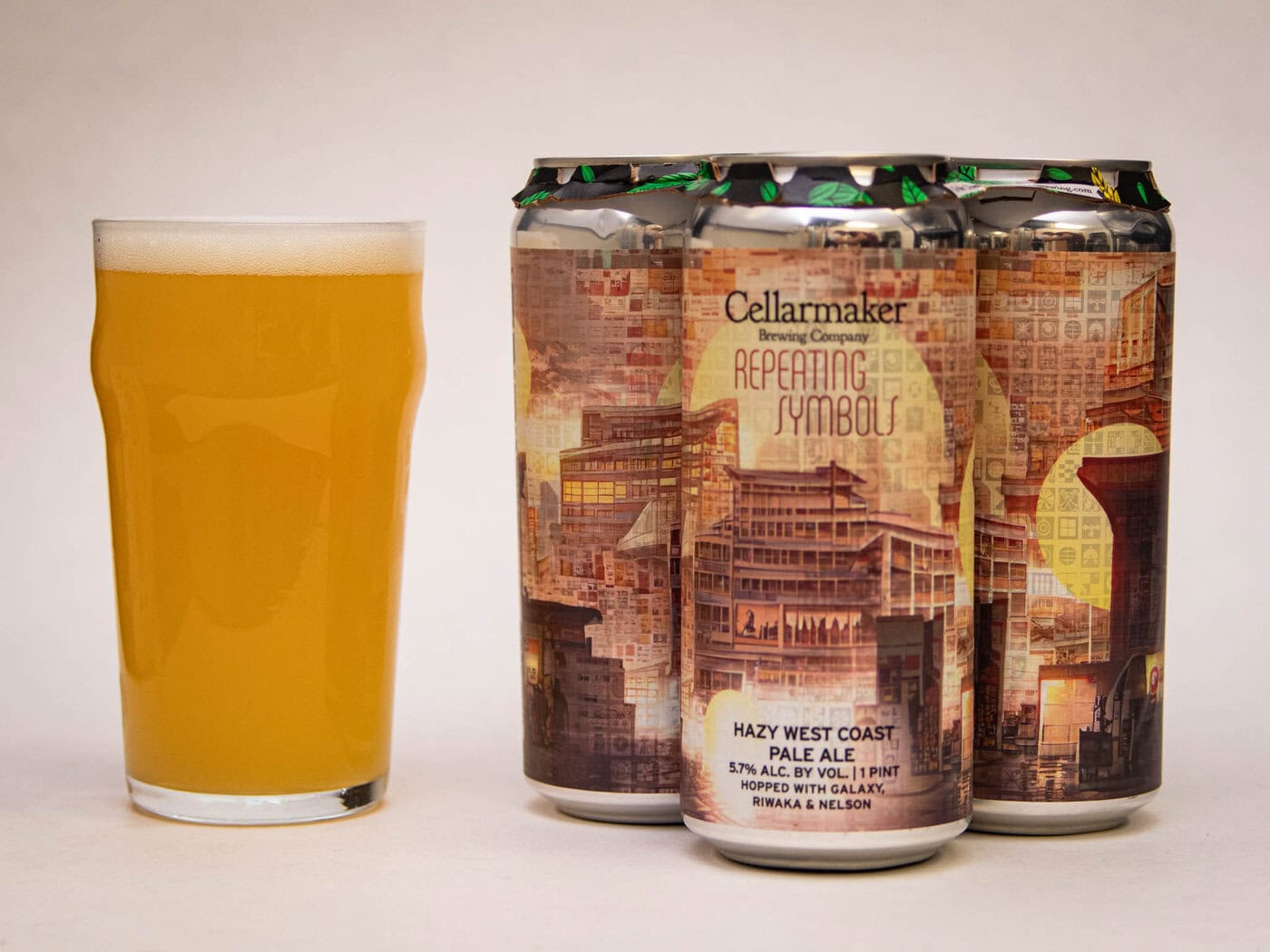 A Pint Glass Filled With Hazy, Pale Yellow Beer Sits Next To Three Cans Of Cellarmaker Brewing Company'S Repeating Symbols Hazy West Coast Pale Ale, Featuring Detailed Cityscape Artwork On The Labels.