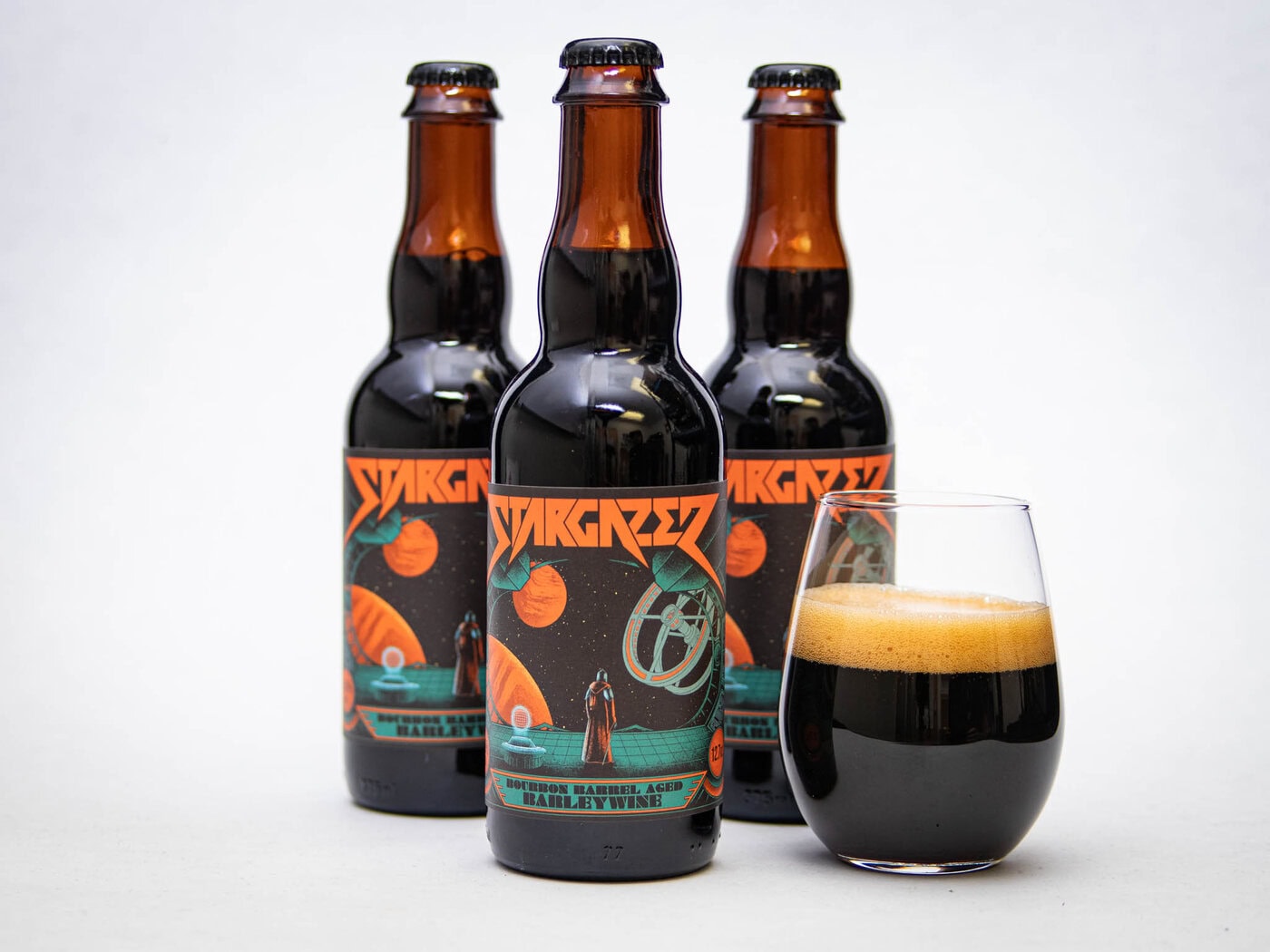 Three Bottles Of Stargazer Beer With Colorful, Sci-Fi Themed Labels Stand Behind A Glass Filled With Dark Beer And A Creamy Foam Head, All Set Against A Plain White Background.