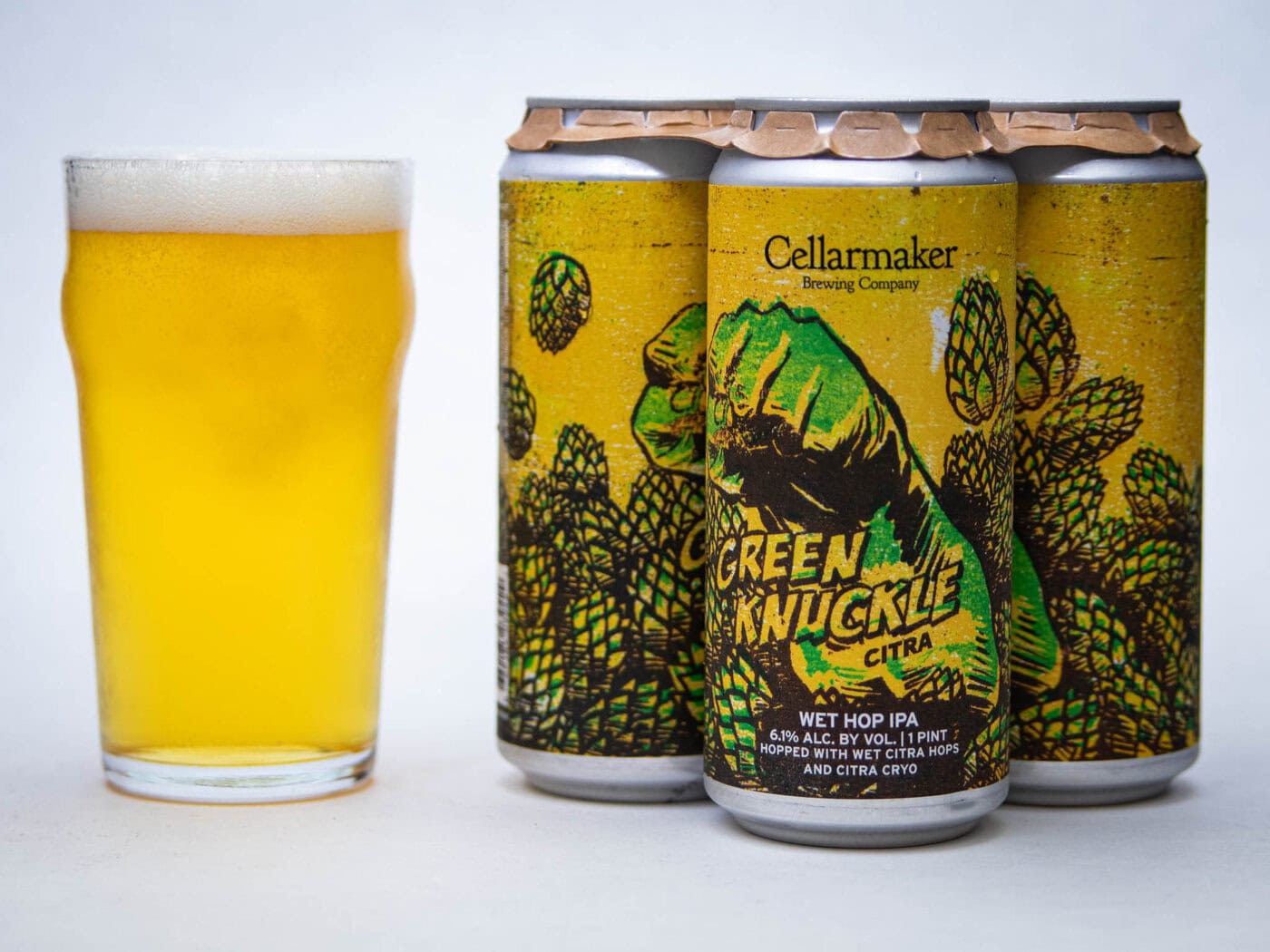 A Pint Glass Of Yellow Beer Sits Next To Three Cans Of Cellarmaker Brewing Company’s Green Knuckle Citra Wet Hop Ipa, Featuring Artwork Of A Green Fist And Hop Plants On A Bright Yellow Background.