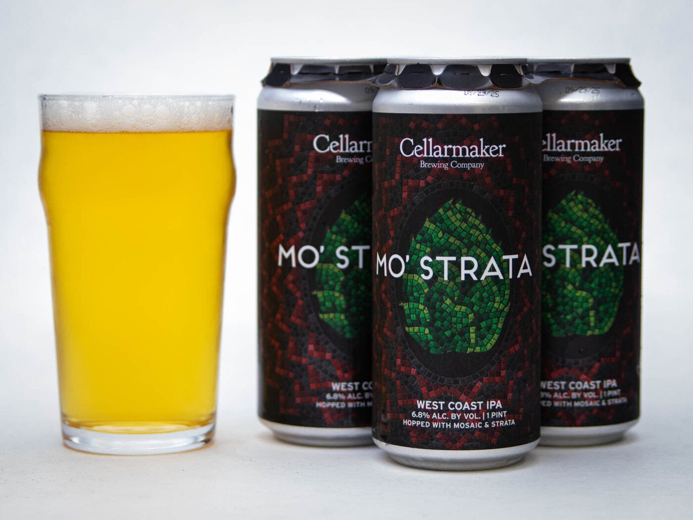 A Pint Glass Of Hazy Golden Beer Sits Next To Three Cans Of Cellarmaker Brewing Company’s Mo’ Strata West Coast Ipa, Featuring A Dark Label With Green Mosaic Artwork.