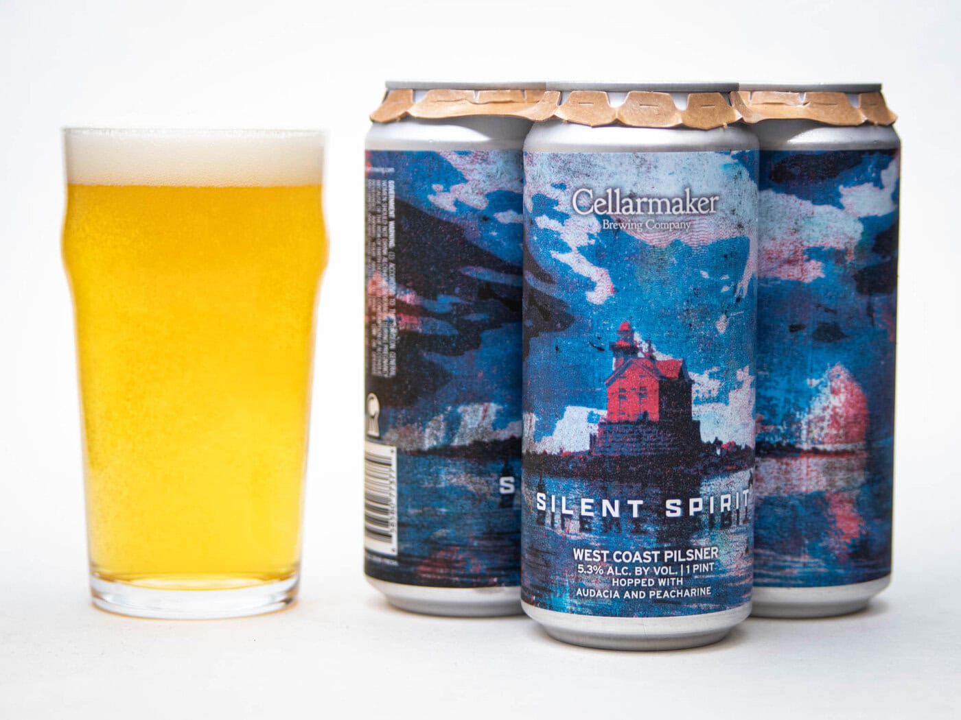 A Full Pint Glass Of Light Golden Beer Sits Next To Three Cans Labeled &Amp;Quot;Cellarmaker Silent Spirit&Amp;Quot; With Blue And Red Artwork Featuring A Lighthouse And Ocean Scene. The Cans Are 16 Oz West Coast Pilsner.