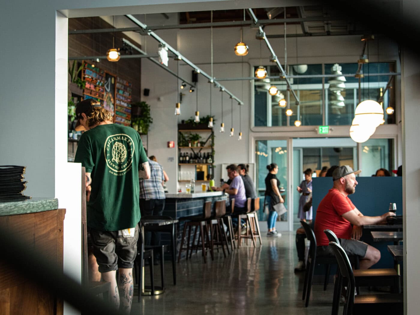 A Casual, Modern Bar With Hanging Lights, People Sitting At Tables And The Bar, And A Man In A Green T-Shirt Standing Near The Counter. The Atmosphere Appears Lively And Relaxed.