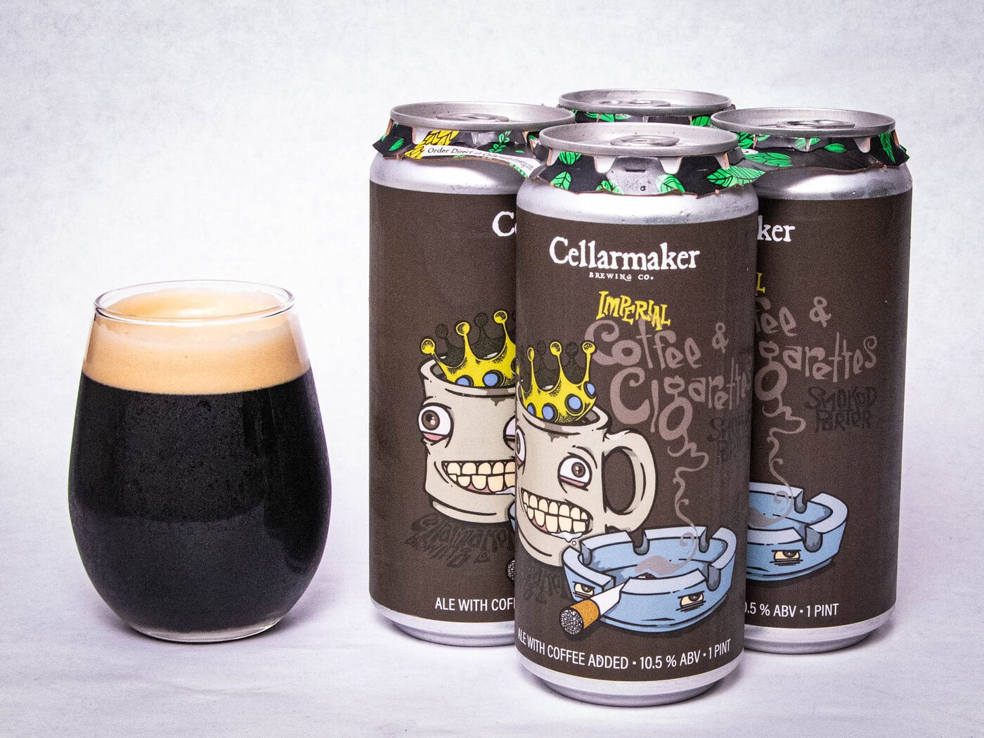 A Glass Of Dark Beer Sits Beside Four Cans Of Imperial Coffee &Amp;Amp; Cigarettes - Coffee Porter, Each Labeled With Cartoon Mugs Wearing Crowns And A Blue Ashtray.