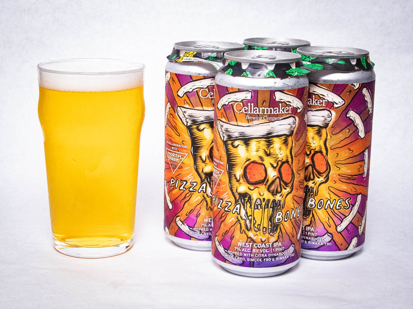 A Pint Glass Of Hazy Golden Beer Sits Next To A Four-Pack Of Colorful Cellarmaker &Amp;Quot;Pizza Bones&Amp;Quot; West Coast Ipa Cans, Featuring A Graphic Of A Skull With Pizza Slices And Bones On The Label.