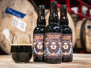 Three bottles of "Steel Your Dreams - BA Barleywine" with illustrated labels stand on a wooden surface beside a glass of dark beer; large barrels appear blurred in the background.