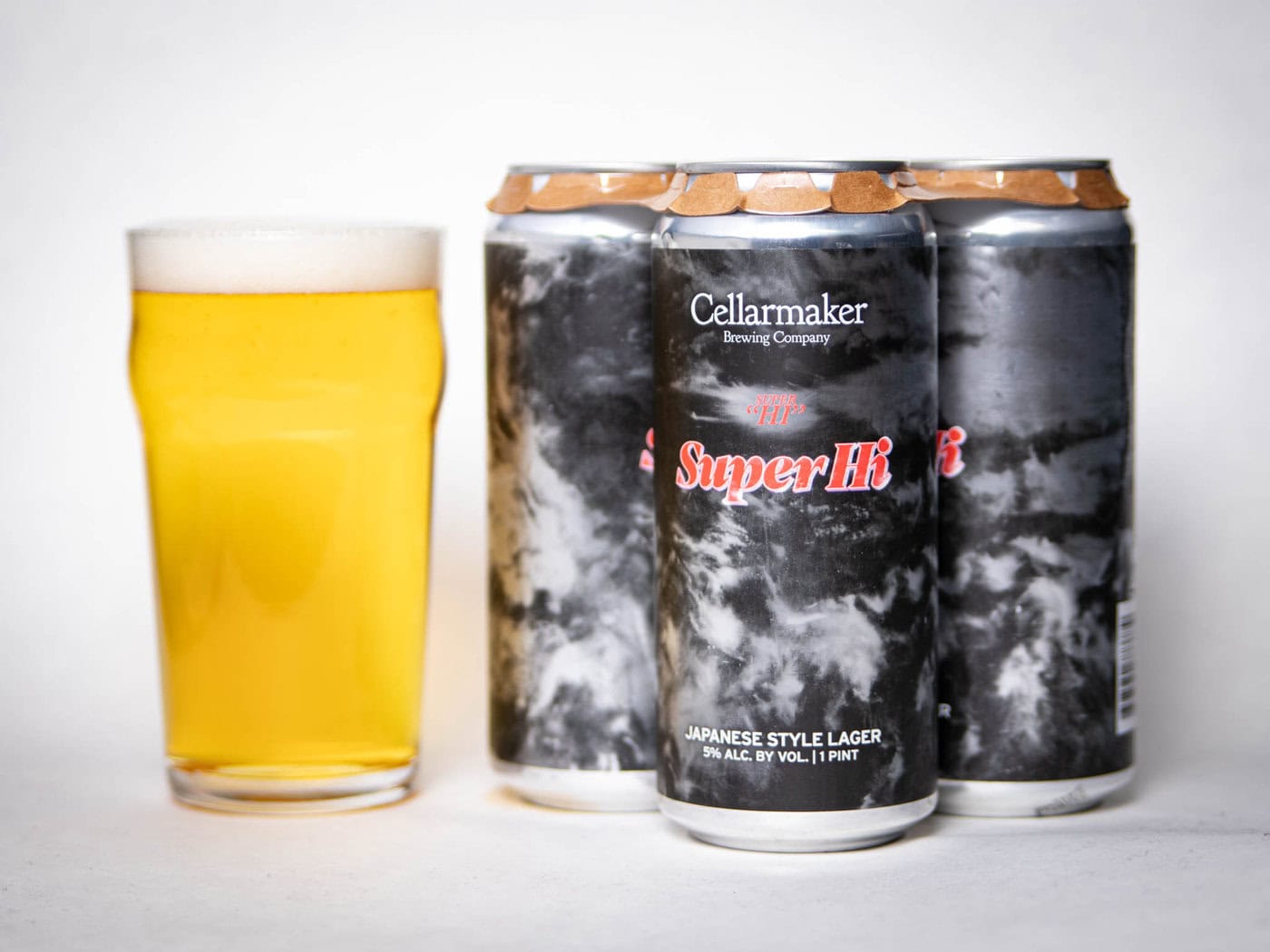 A Pint Glass Filled With Pale Yellow Beer Sits Next To Three Cans Labeled &Amp;Quot;Cellarmaker Super Hi! Japanese Style Lager&Amp;Quot; With A Dark, Cloudy Design On The Cans.