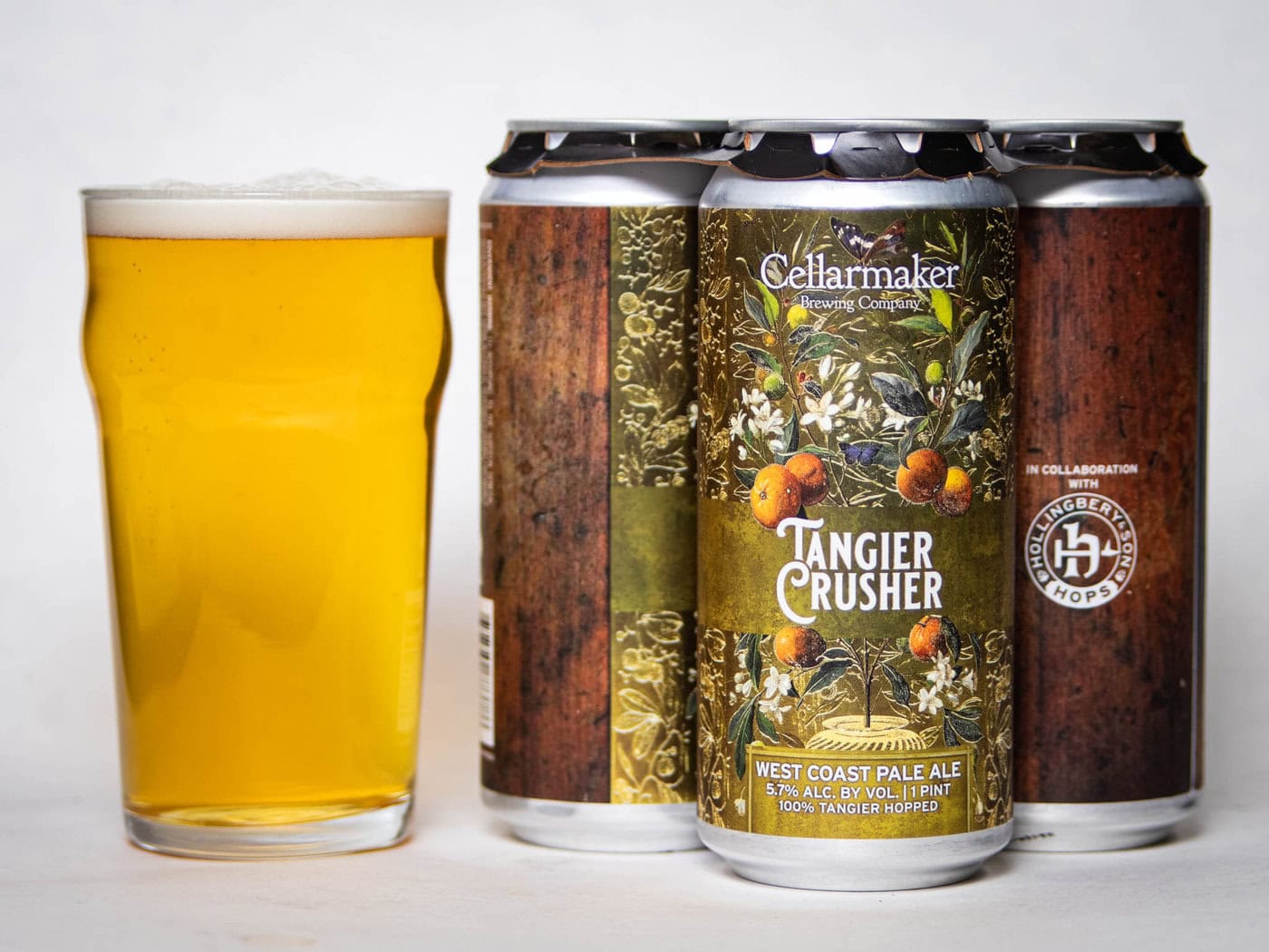 A Pint Glass Filled With Hazy Golden Beer Stands Beside Three Cans Of Cellarmaker Brewing Co.'S Tangier Crusher West Coast Pale Ale, Featuring Floral And Fruit Artwork On The Labels.
