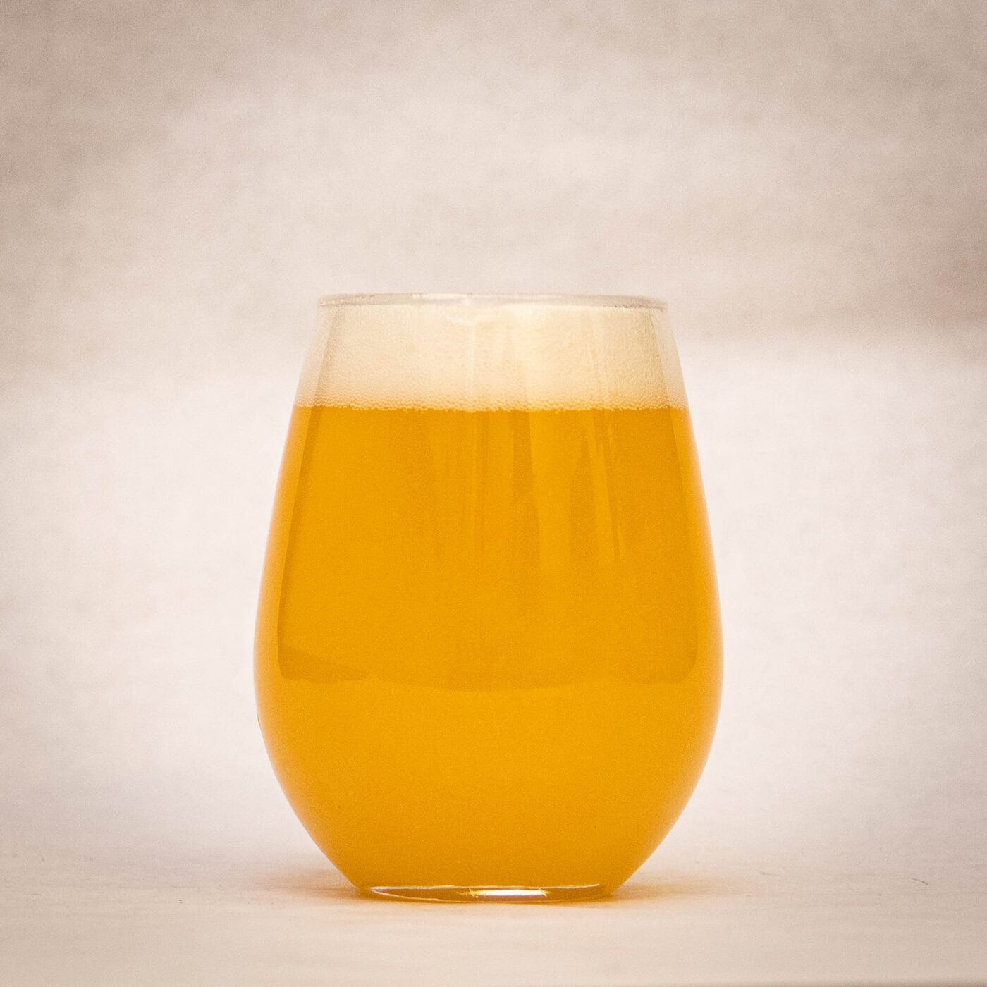 A Stemless Glass Filled With A Hazy, Golden-Yellow Beverage Sits On A Plain Surface Against A Light, Neutral Background. The Drink Has A Foamy Layer At The Top.