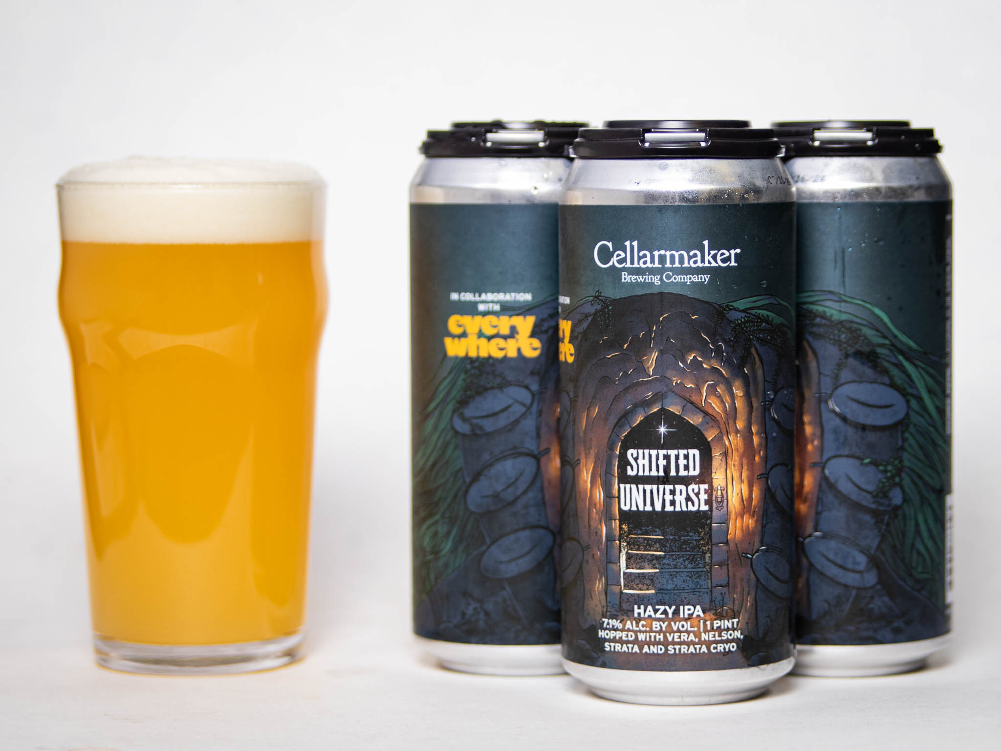 A Pint Glass Of Hazy, Golden Beer Sits Next To Four Cans Labeled &Amp;Quot;Shifted Universe Hazy Ipa&Amp;Quot; By Cellarmaker Brewing Company. The Cans Feature Dark, Illustrated Tunnel Artwork With Glowing Light At The Center.