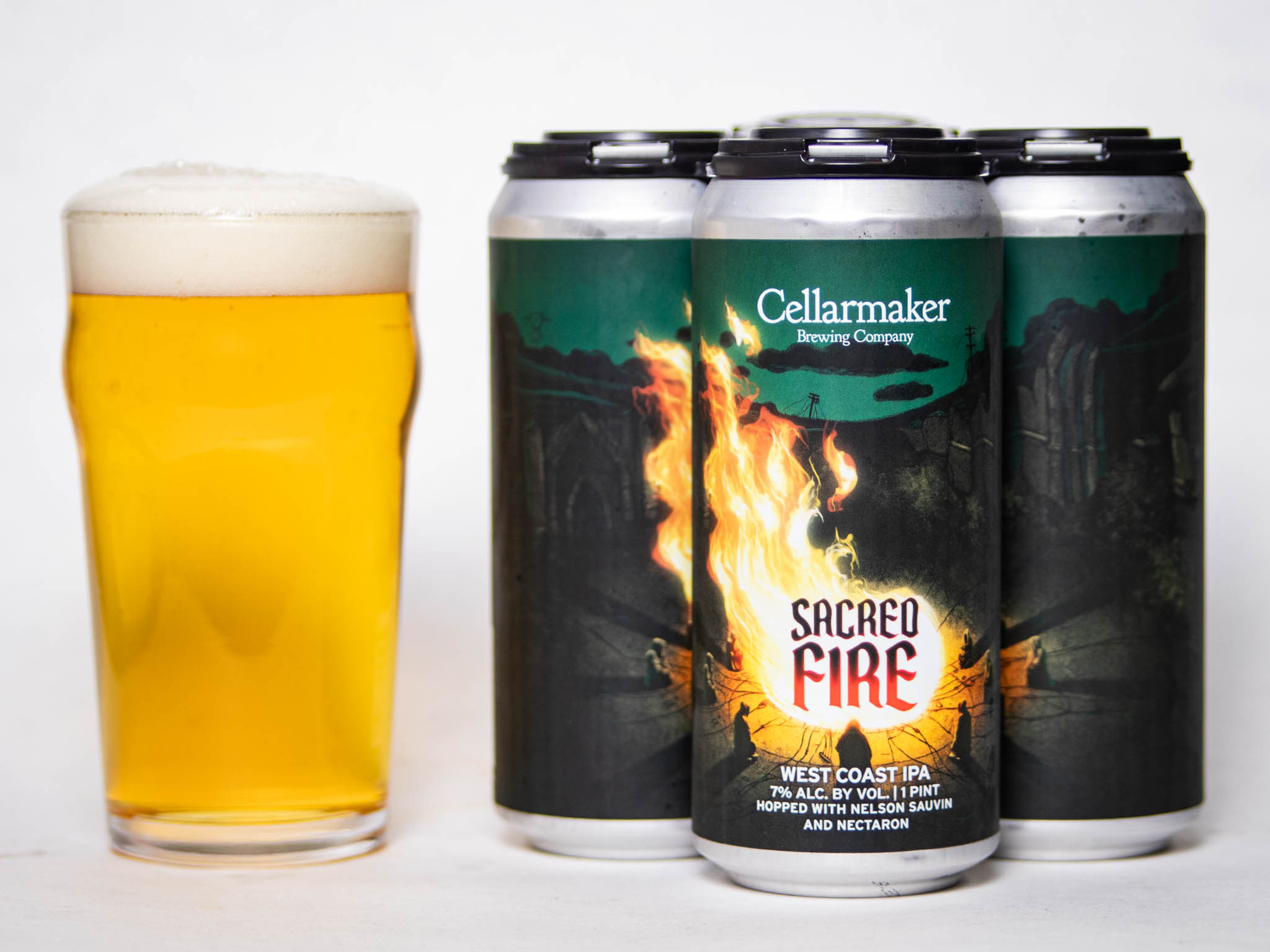 A Pint Glass Filled With Foamy Beer Sits Next To Four Cans Of Cellarmaker Brewing Company'S Sacred Fire West Coast Ipa, Featuring A Fiery Label Design With Dark, Mountainous Scenery In The Background.
